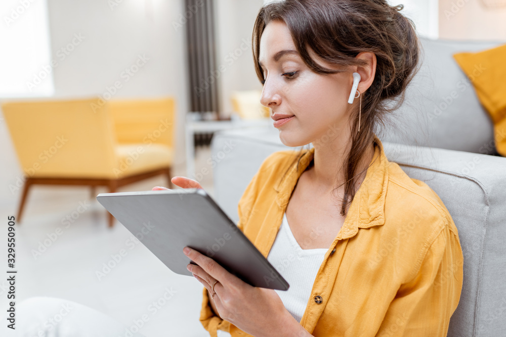 一位年轻开朗的女士，一边放松地坐着，一边使用无线耳机在数字平板电脑上工作
