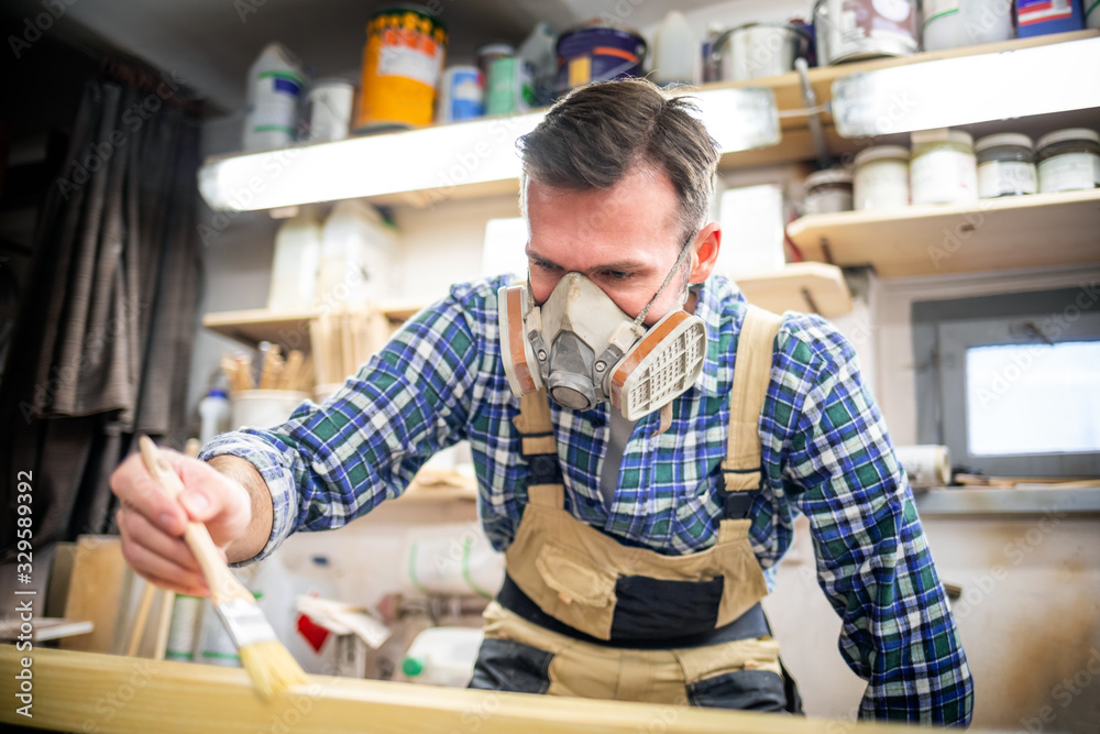 Carpenter with mask applies paint using paintbrush in carpentry workshop