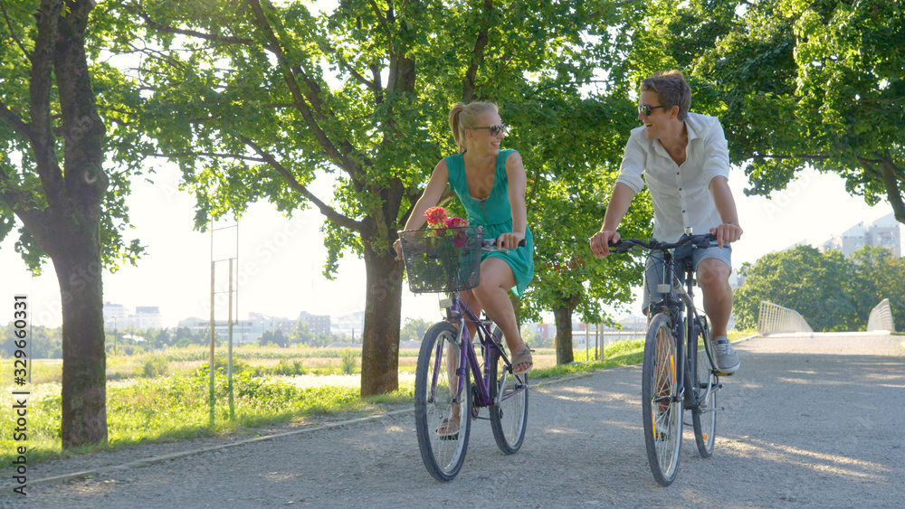 Young Caucasian couple smiles while riding bicycles down a scenic green avenue.