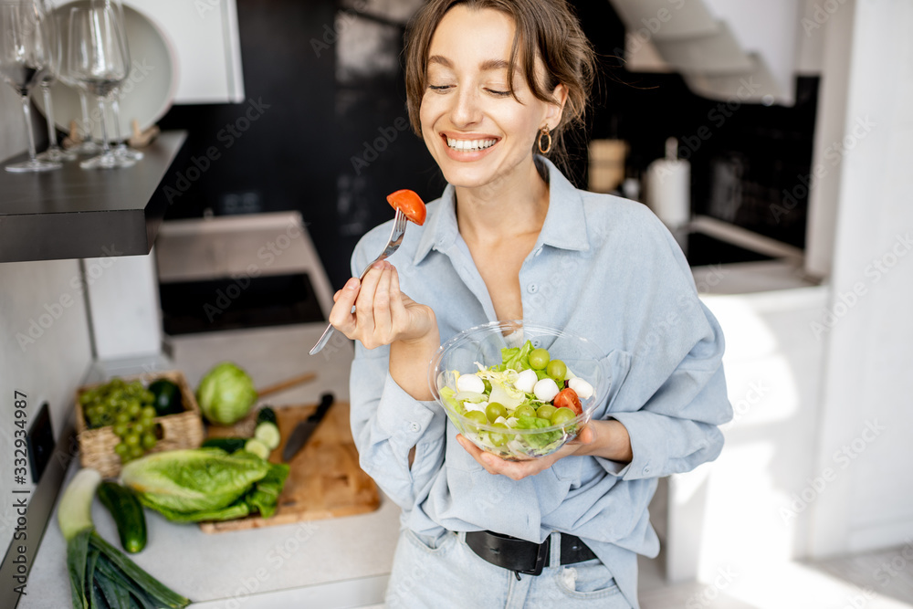 一个年轻快乐的女人在家里的厨房里吃健康沙拉的画像。健康饮食，