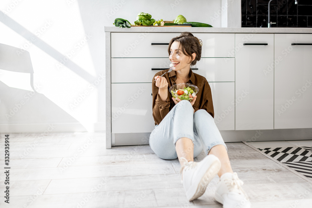 Young woman sitting on the white kitchen floor, eating salad while relaxing at home. Healthy eating,