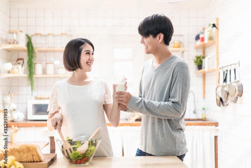亚洲新婚夫妇一家站在温暖的厨房里。孕妇和丈夫一起拿着一杯牛奶