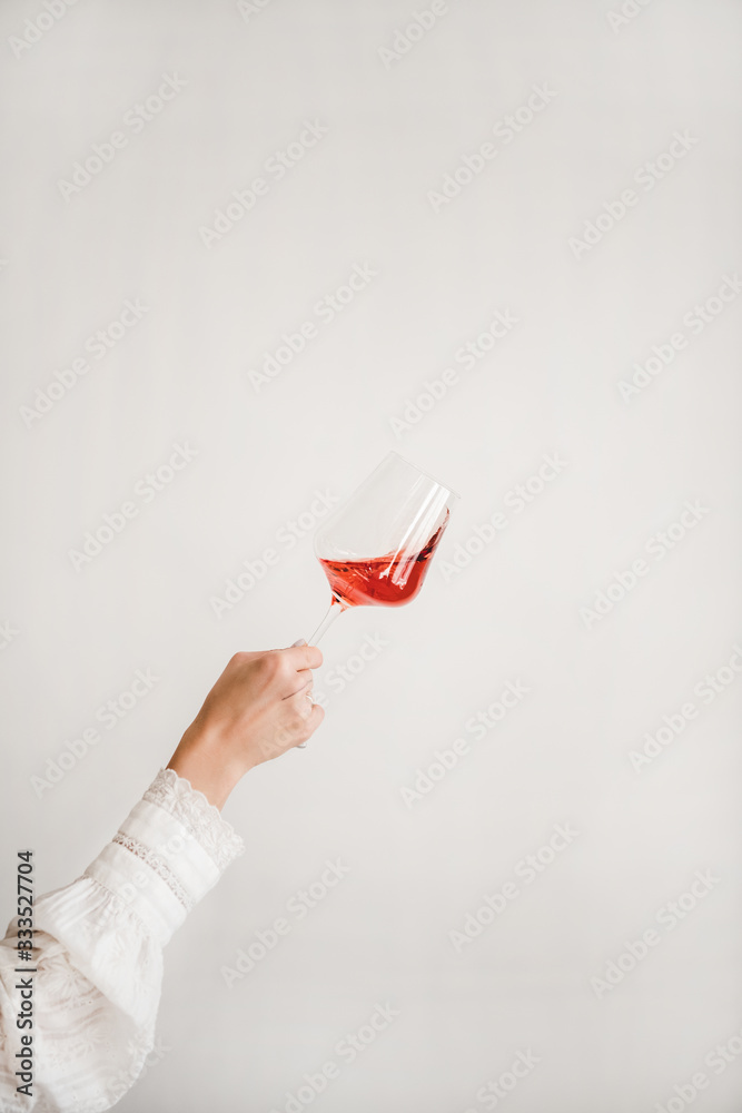 Womans hand in white shirt holding and turning glass of rose wine during tasting over white wall bac