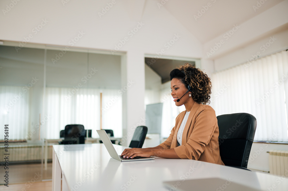 微笑的混血女性在明亮的办公室里工作，使用笔记本电脑和耳机，肖像。