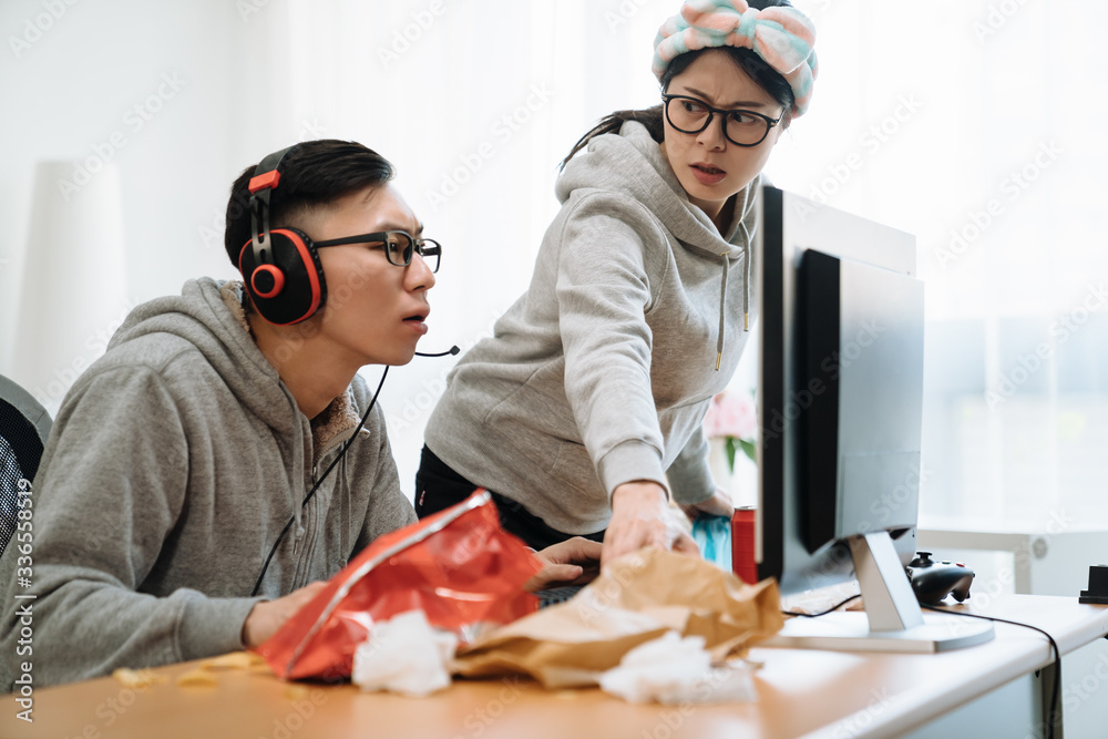 戴着耳机的韩国男子在电脑上玩网络游戏，而愤怒的妻子站着盯着嗨