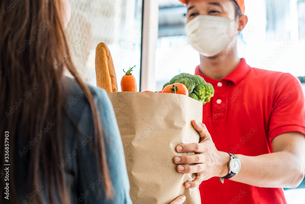 亚洲送货员戴着口罩，穿着红色制服，处理食品、水果和蔬菜的袋子。