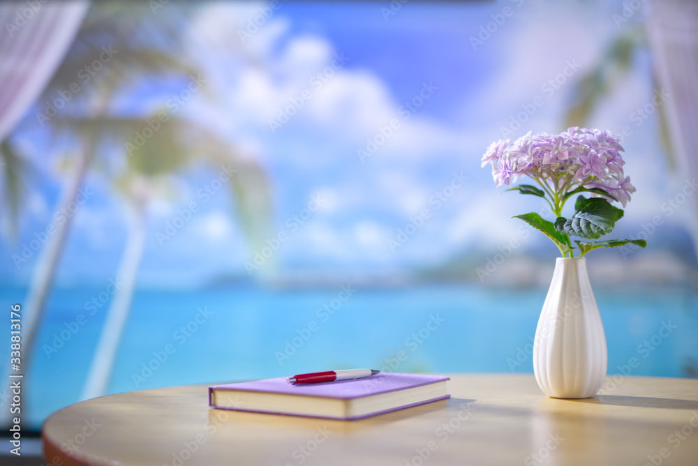 海滨别墅内部木制桌子，配有笔记本、钢笔和一瓶紫色的花。美丽的blurr