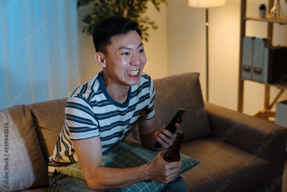 young asian korean man watching sports competition and drinking beer in comfort couch in home living