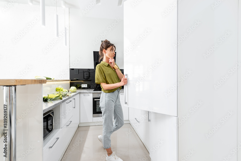 女人在家里的现代厨房里烹饪健康食品，看着冰箱，尽情享受
