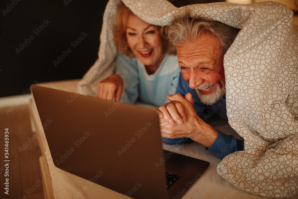 一对面带微笑的老年夫妇躺在床上，盖着毯子进行视频通话。