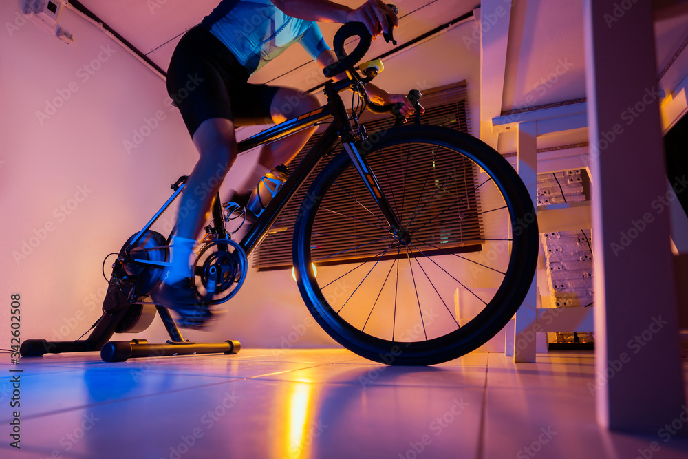 亚洲男子骑着机器教练自行车，晚上在家锻炼。他玩网上自行车g