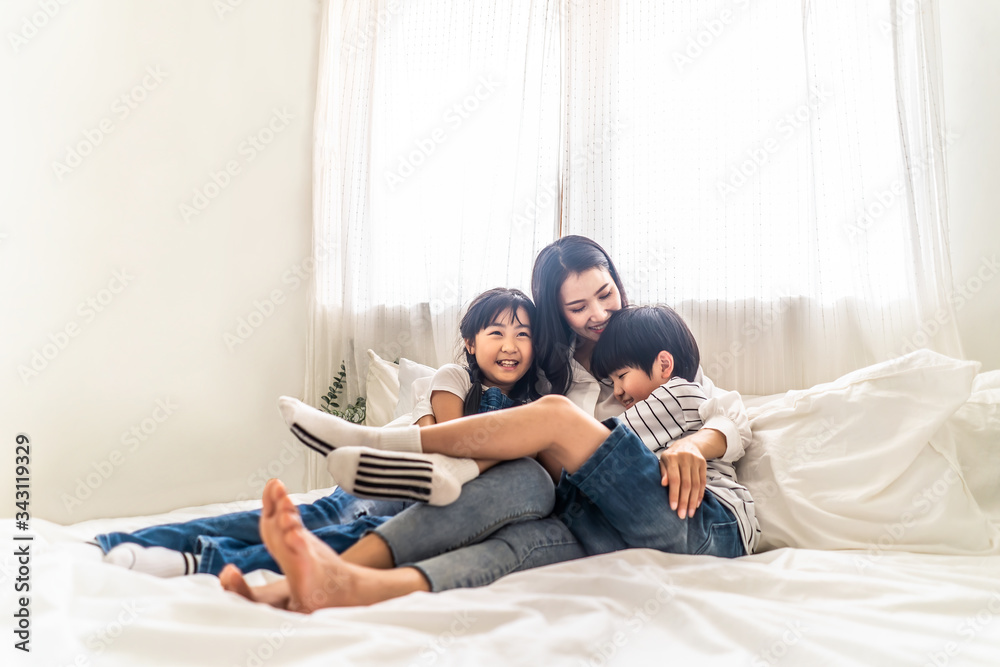 亚洲家庭的母亲、儿子和女儿幸福地坐在白色的床上，在卧室里微笑。年轻的博