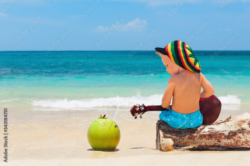 Little baby in rasta hat play reggae music on Hawaiian ukulele, enjoy relaxing on ocean beach. Kids 