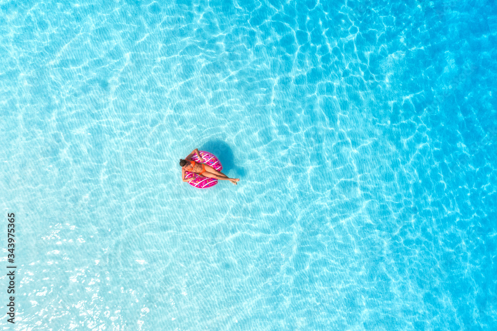 日落时，一名年轻女子带着粉色甜甜圈在透明的蓝色大海中游泳的鸟瞰图