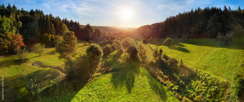 Aerial landscape panorama after sunrise: gorgeous scenery with the sun in the blue sky, trees on gre