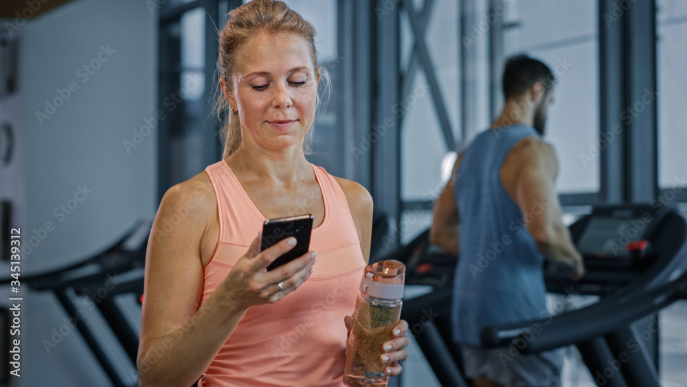 Beautiful Athletic Woman Uses Smartphone Walks Through Fitness Club Gym. Using Social Media, Posting