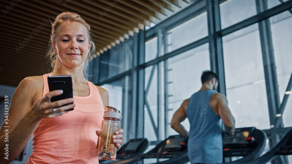 美丽的运动女性使用智能手机在健身俱乐部健身房散步。使用社交媒体，发帖