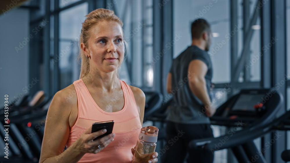 美丽的运动女性使用智能手机在健身俱乐部健身房散步。使用社交媒体，发帖