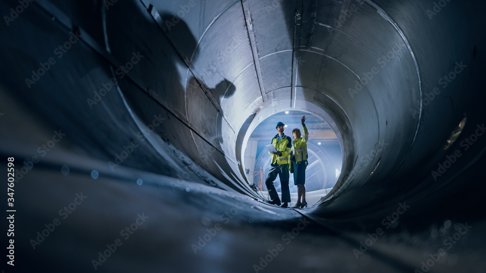 Two Heavy Industry Engineers Walking Inside Pipe, Use Laptop, Have Discussion, Checking Design. Cons