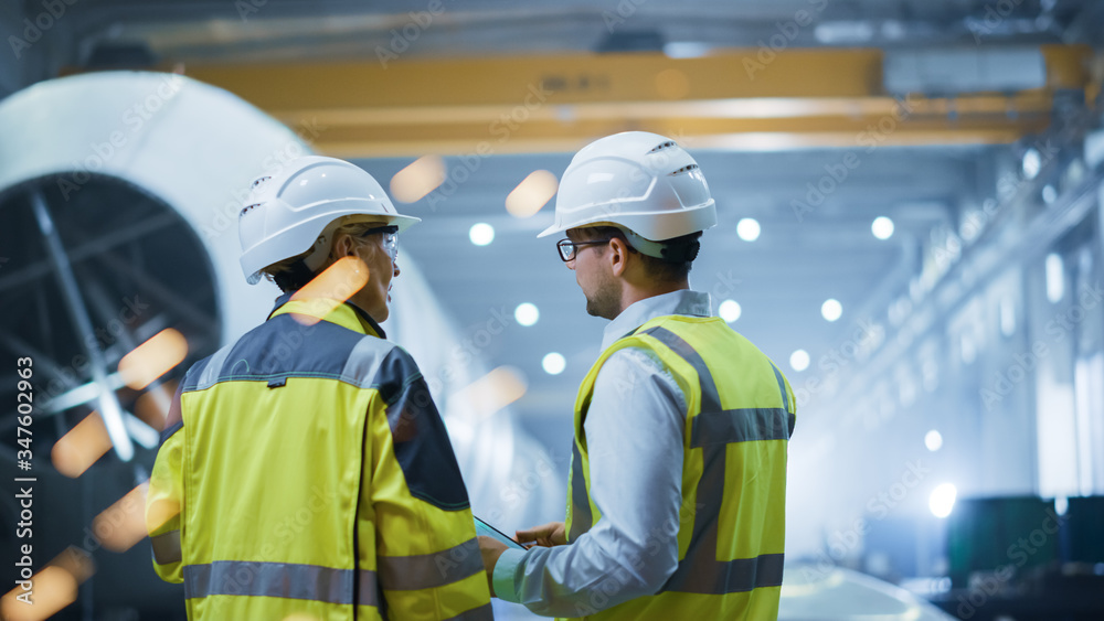 Two Heavy Industry Engineers Stand in Pipe Manufacturing Factory, Use Digital Tablet Computer, Have 