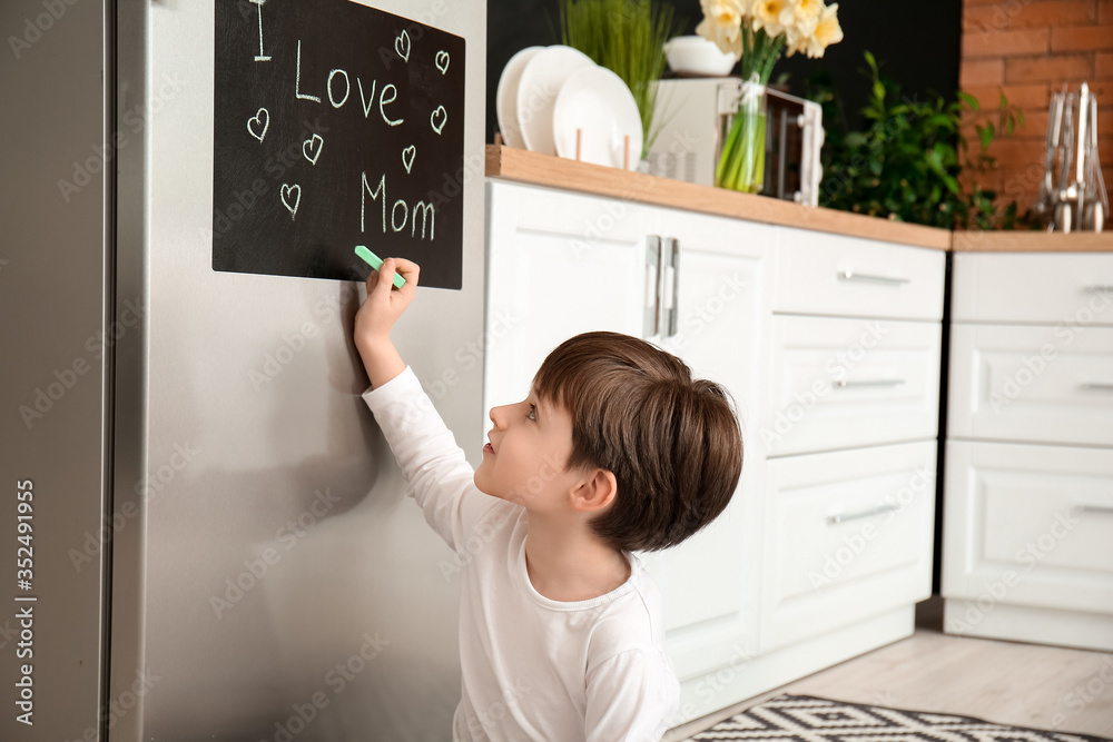 Little boy writing text I LOVE MOM on chalkboard in kitchen