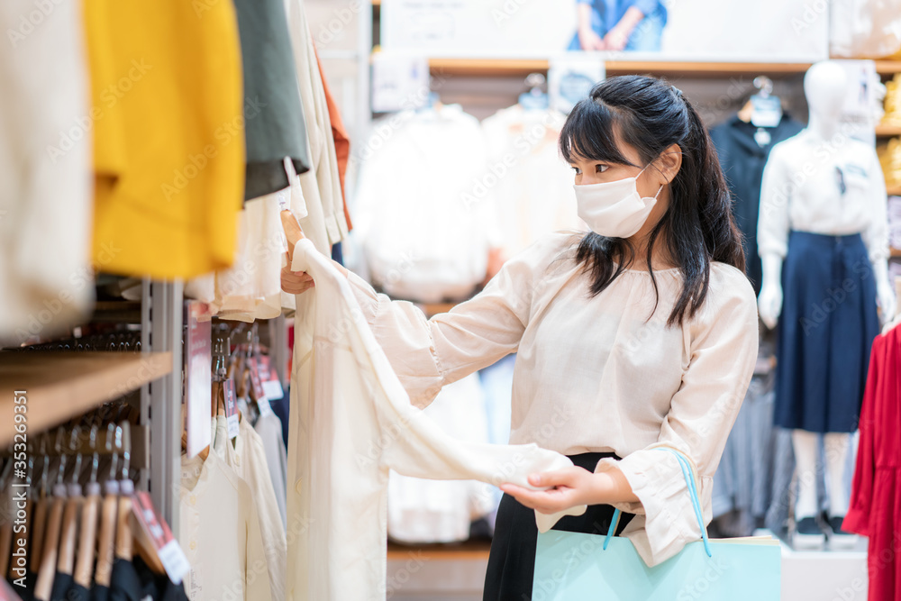 一名亚洲女子在购物中心用购物袋选择衬衫时戴着口罩