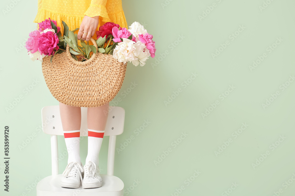 Young woman with beautiful peony flowers in bag standing on chair against color background