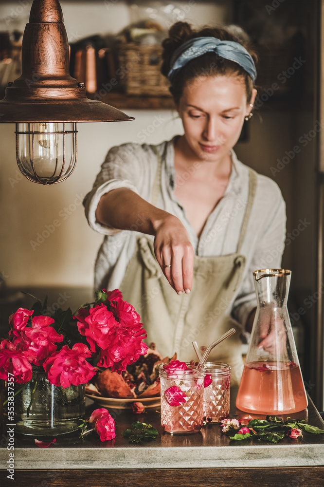 一位穿着亚麻围裙的年轻女士在混凝土套装上的玻璃杯里加冰，为玫瑰柠檬水添加配料