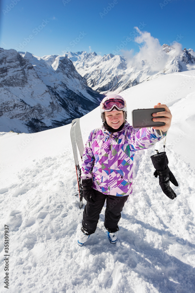 一个女孩戴着滑雪服眼镜，戴着带山峰的头盔，在冬天自拍并微笑的俯视图cl