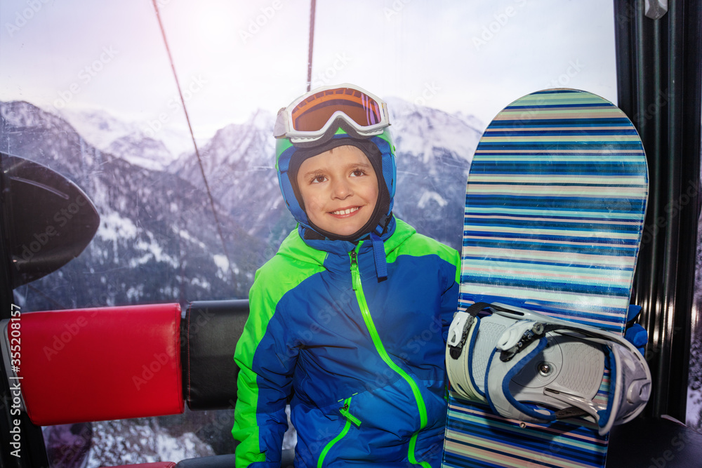 Little boy sit with snowboard and happy smile in the ski lift cabin on winter resort