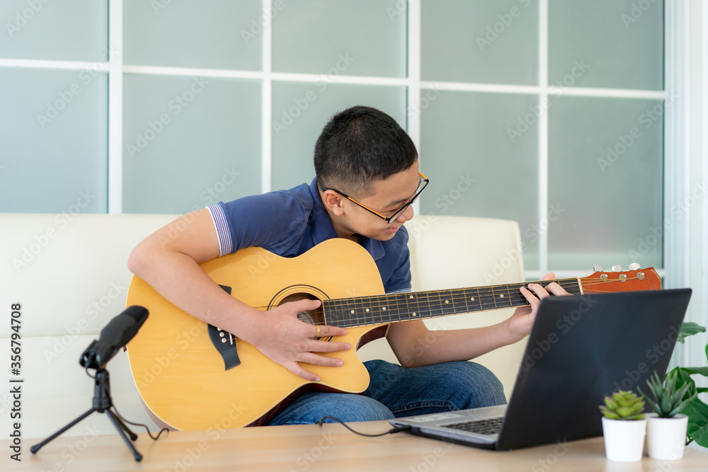 亚洲男孩在家练习时弹原声吉他并在笔记本电脑上观看在线课程。Asi