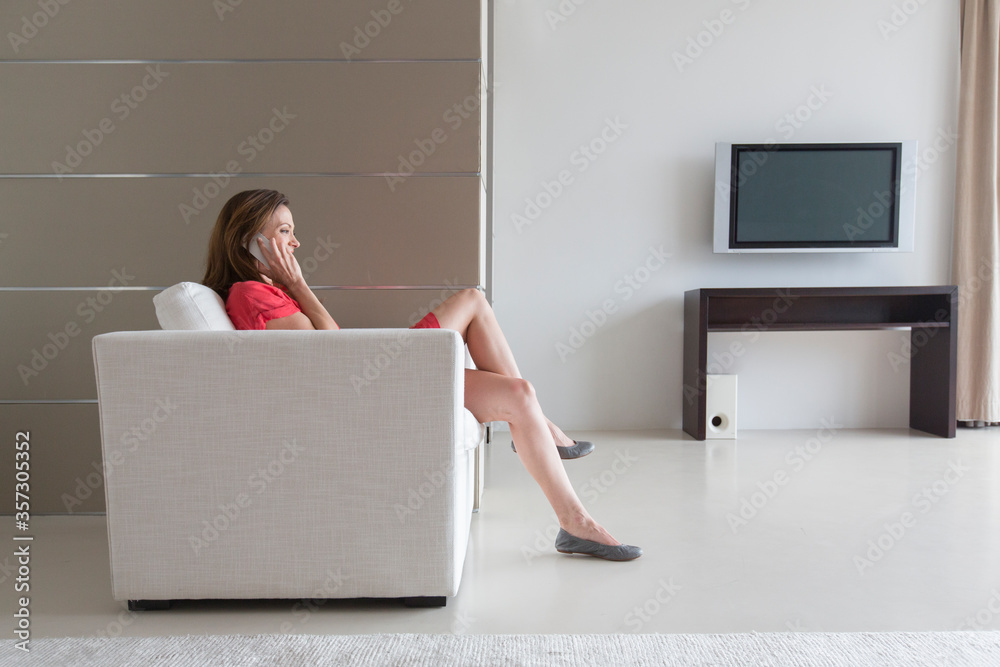 Woman in red dress sitting in armchair talking on phone in modern living room