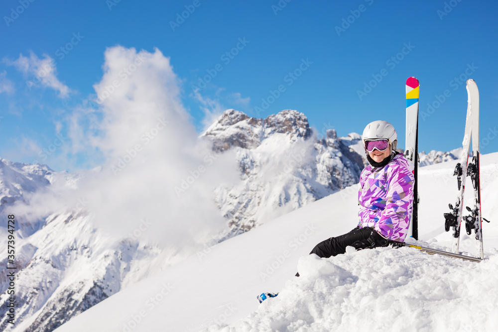 穿着滑雪服、戴着头盔和口罩的快乐女孩坐在山顶的雪地里看着镜头