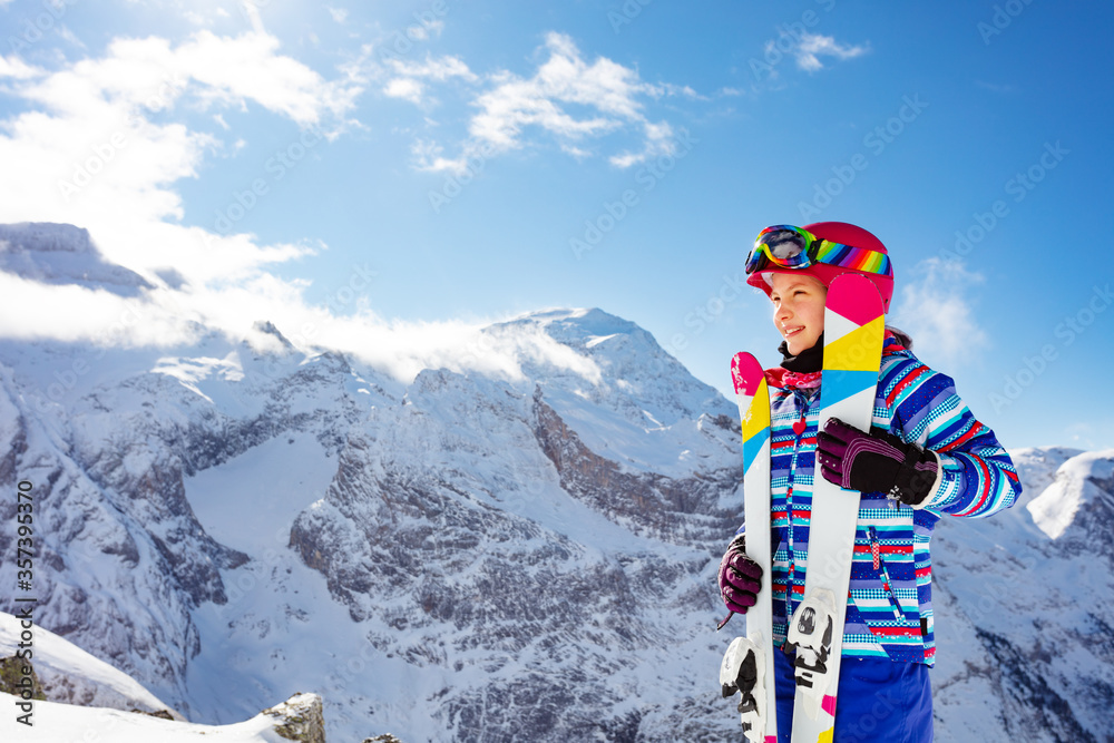 戴着粉色头盔和彩色滑雪板，摘下护目镜，在高山上微笑的可爱女孩的肖像