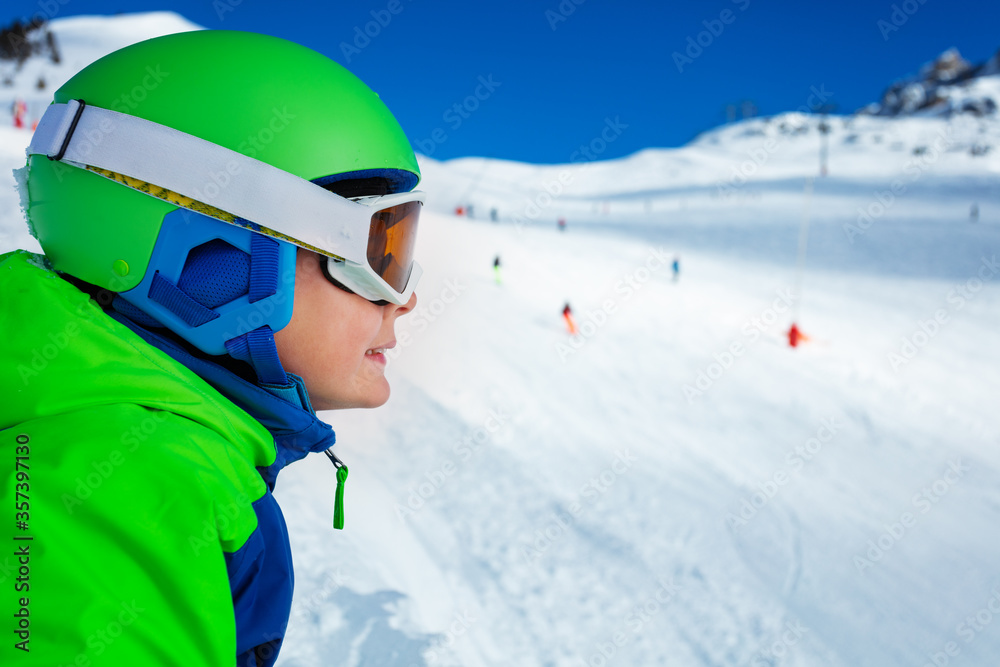 一个戴着口罩和头盔的男孩在滑雪板上的近照，从滑雪道上看向一边