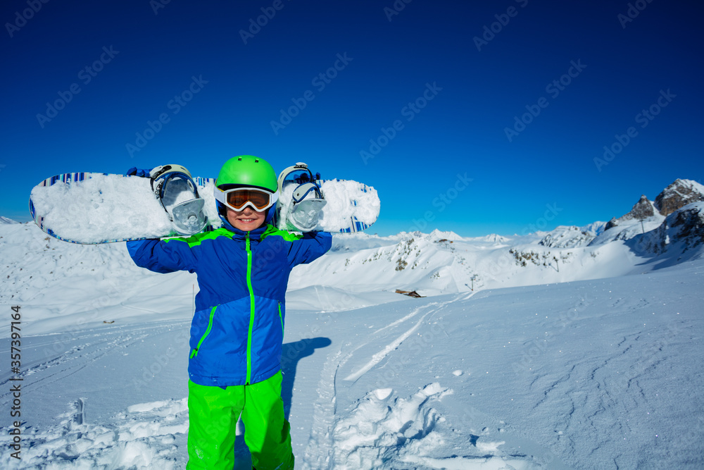 小男孩戴着头盔，带着滑雪板站在山脉的雪地里