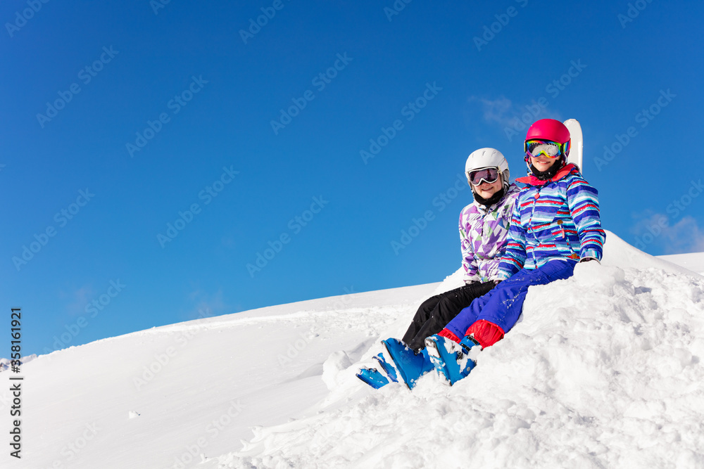 两个可爱的女孩穿着天空服装，戴着彩色口罩和眼镜，坐在蓝色背景下的雪地上