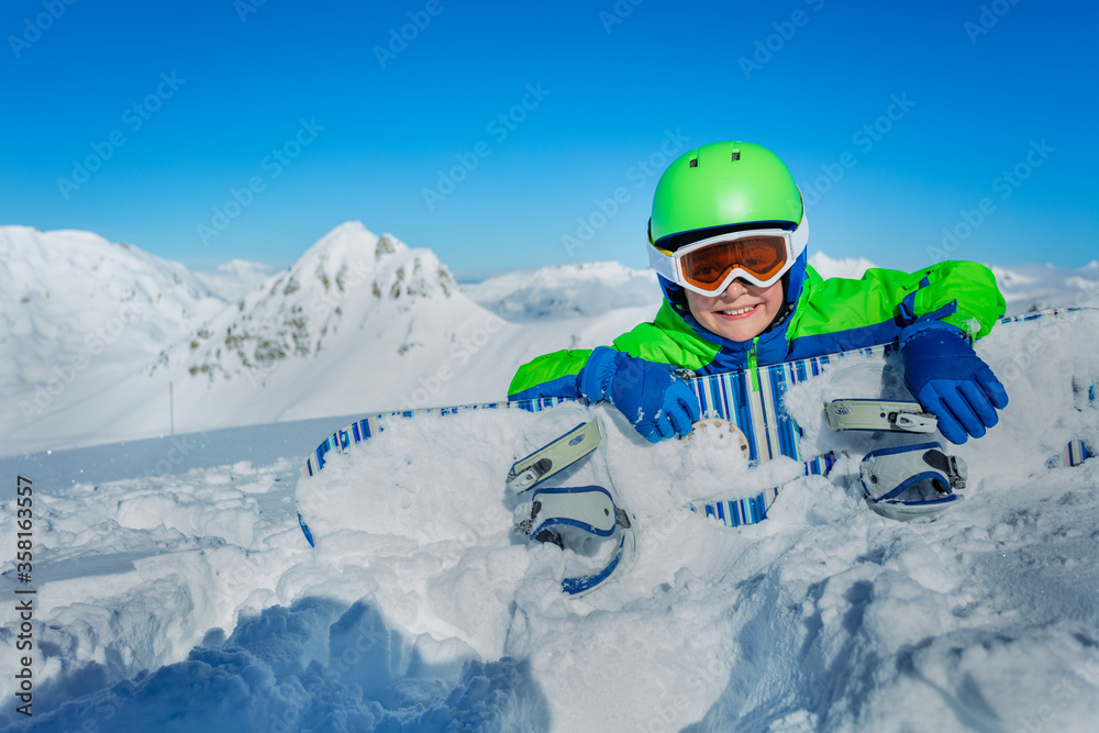 小男孩戴着头盔和滑雪谷歌眼镜，躺在雪地里滑雪
