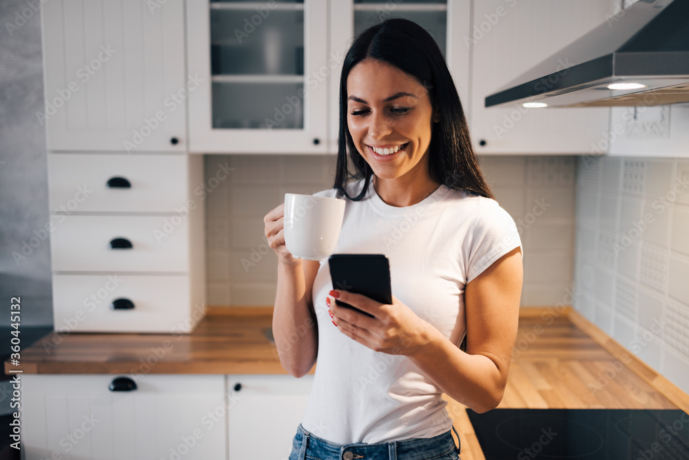 一个可爱快乐的女人喝早茶并使用智能手机的肖像。