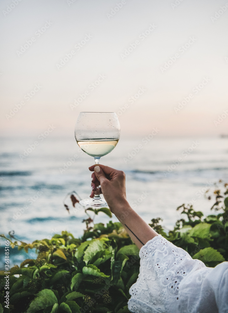 Womans hand in white lace dress sleeve holding glass of white wine with beautiful sunset colors and 