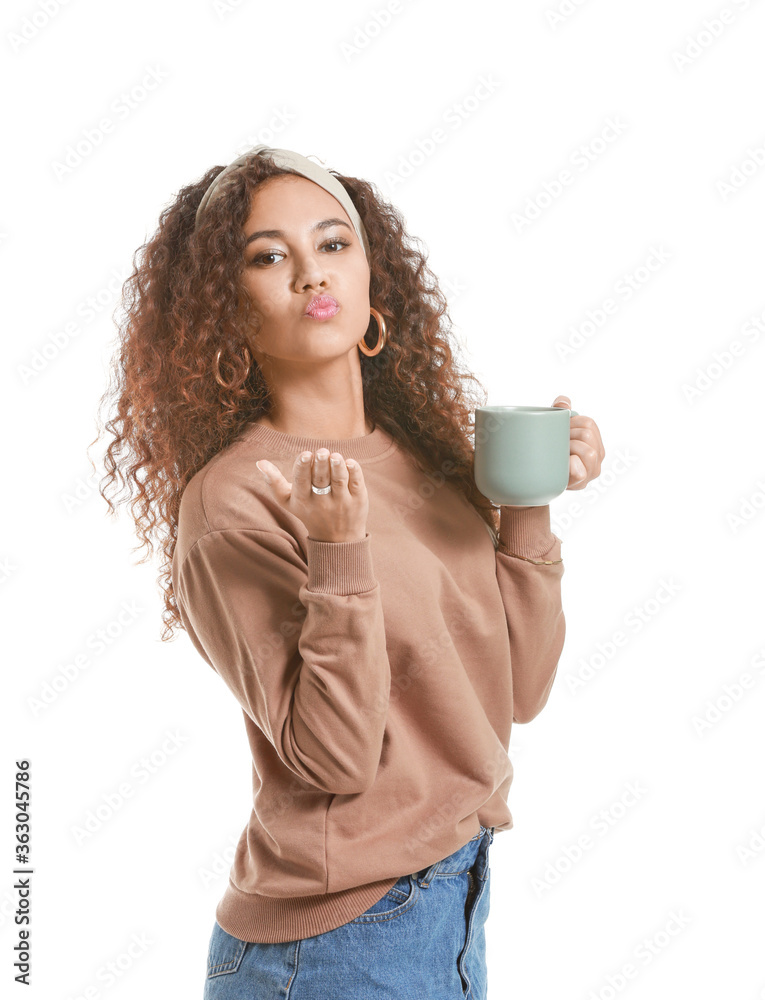 Beautiful young African-American woman with hot tea blowing kiss on white background