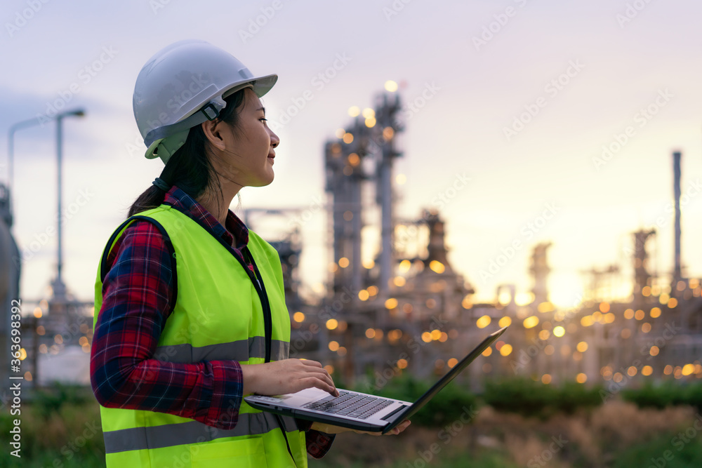亚洲女石化工程师在石油和天然气炼油厂里拿着笔记本电脑晚上工作