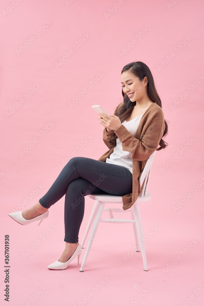 一位满意的休闲亚洲女性坐在粉色连衣裙上的椅子上使用手机的肖像