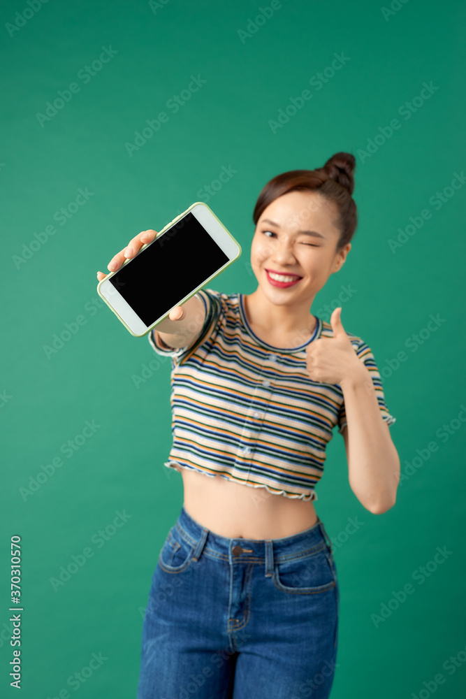 一位微笑的亚洲女性在绿色背景下展示空白屏幕手机的特写。