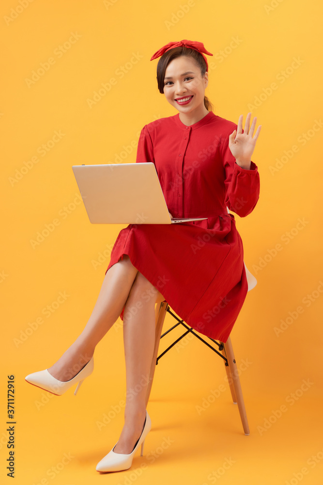 一位漂亮的年轻亚洲女性，她用笔记本电脑，坐在橙色背景下的扶手椅上。