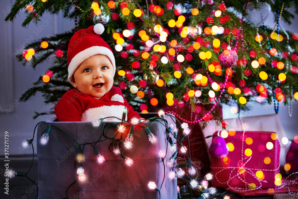 戴着圣诞老人帽子的小可爱男婴，手里拿着发光的灯，坐在圣诞帐篷下的盒子里