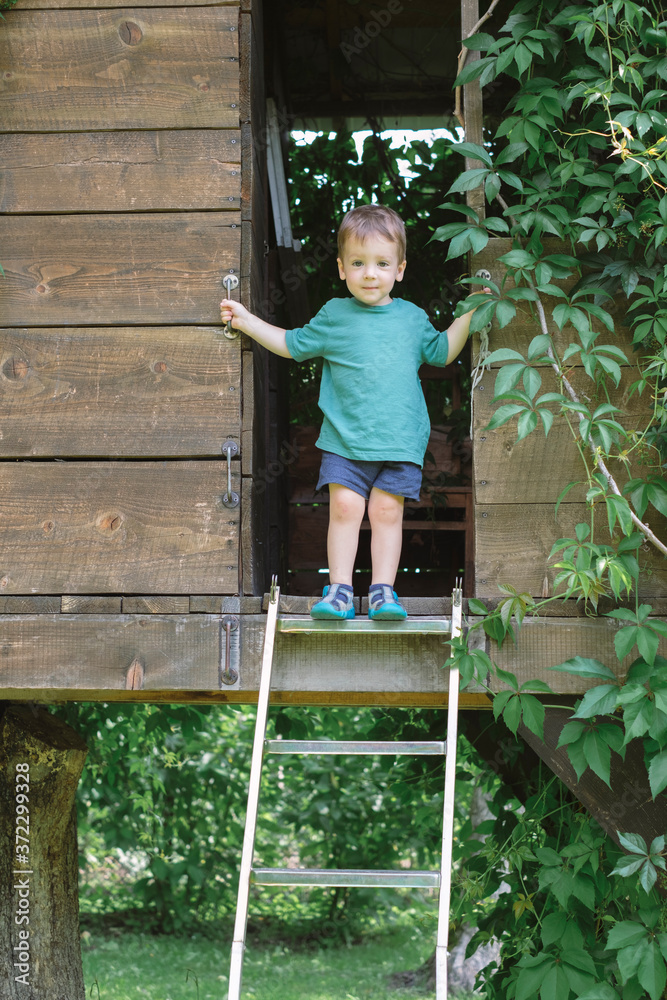 小男孩站在绿色花园树屋的楼梯上。夏天