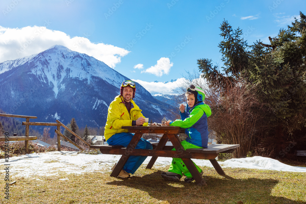 穿着滑雪服的快乐小男孩滑雪后与父亲一起坐在山上欣赏全景，享受午休时光