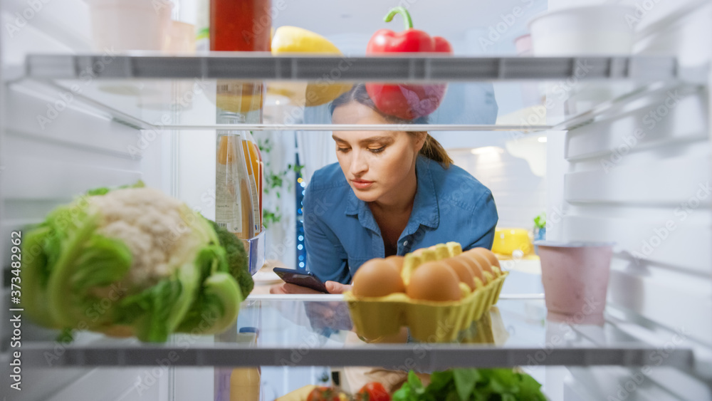 年轻女人打开冰箱门，在智能手机中查看食谱。女人在准备健康餐。Poi