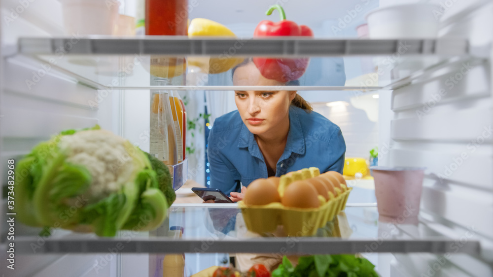 年轻女子打开冰箱门，在智能手机中查看食谱。妇女正在准备健康餐。Poi